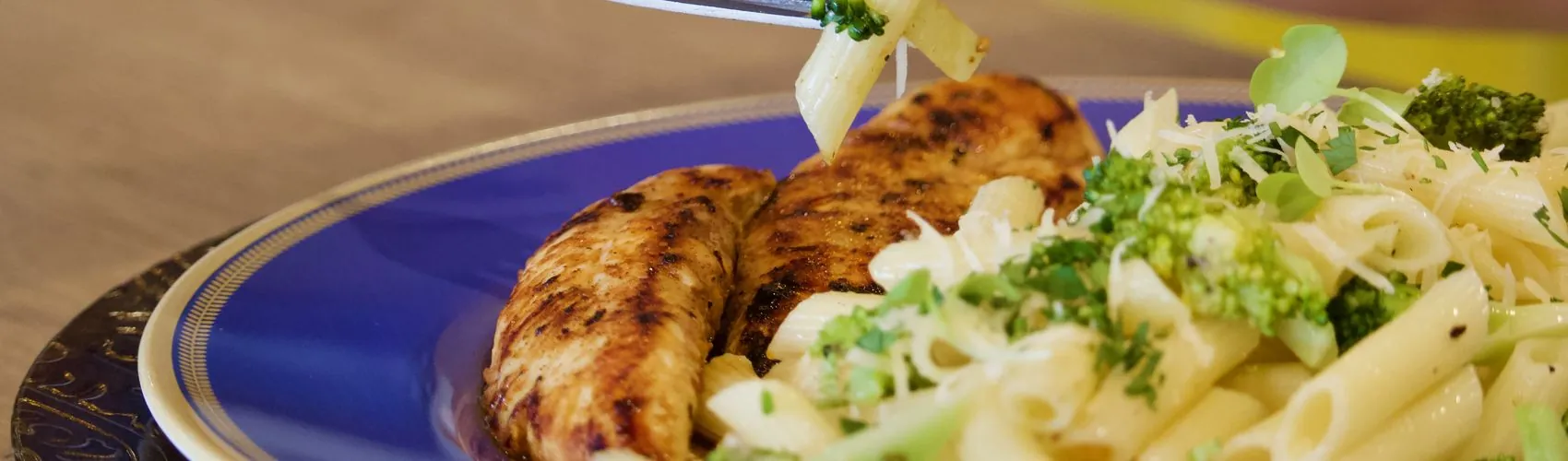 Prato de filé de frango grelhado com penne e brócolis, e um garfo levando uma porção à boca